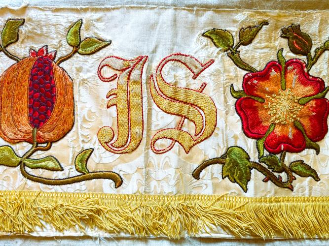 Detail of an embroidered cloth with a piece of fruit and flower shown alongside the word IS