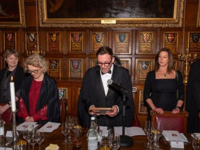 A man in glasses reading in a wood-panelled room with crests and portraits hanging on the wall, with three women on either side