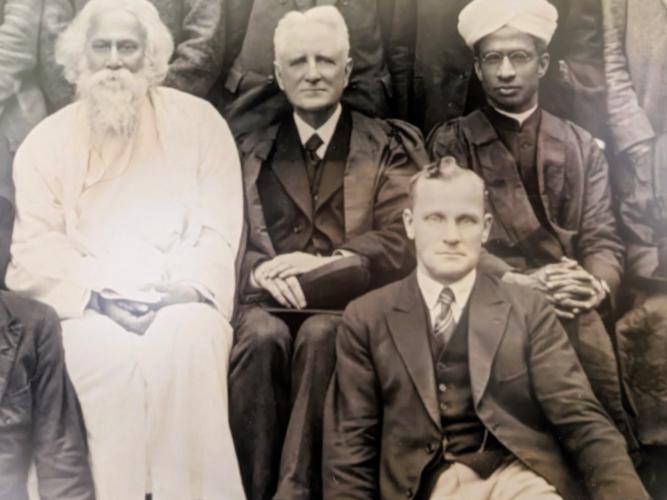 A detail of a photograph showing four figures. Three are seated next to each other on chairs and show from left to right, a man with a white beard and white robes, and two men in suits and academic gowns, one wearing a turban. 