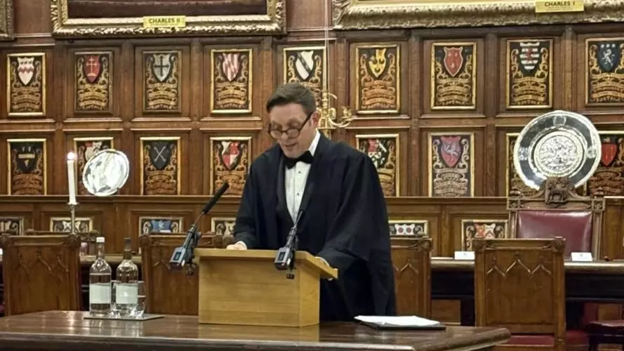 Professor Matthew Weait reading from a lectern in a wood-panelled room with crests and portraits hanging on the wall