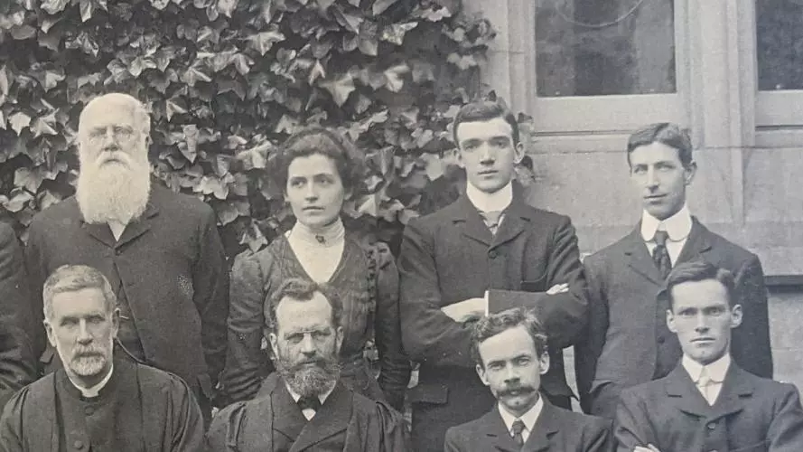 A detail of a black and white photograph showing students posing for a College group picture.  A female figure is standing next to a man with a long white beard.