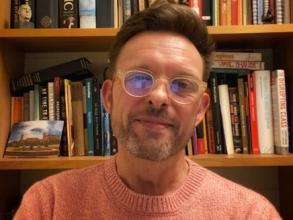 Portrait of Professor Matthew Weait smiling and facing directly ahead. He is wearing a light orange jumper and glasses, and is sitting in front of a bookcase.