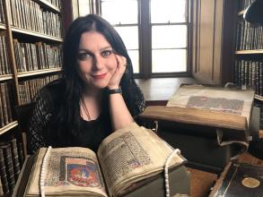 Janina Ramirez posing in front of an old book with bookshelves and windows behind her back.