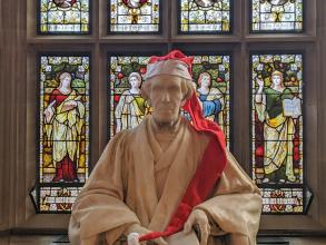 A picture of a marble statue of a man seated in academic robes.  The statue has a long Christmas hat placed on the man's head.
