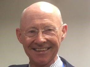 Dr Chris Oneill wearing suit with yellow tie, and a blue lanyard posing in front of a grey background.