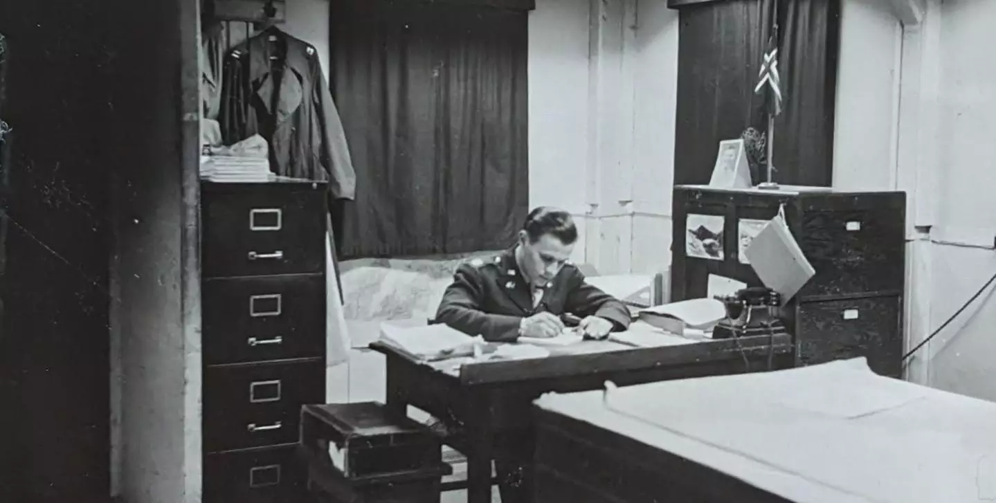 A black and white photograph taken during the second world war of an army staff member working at a desk in front of a large map chest.