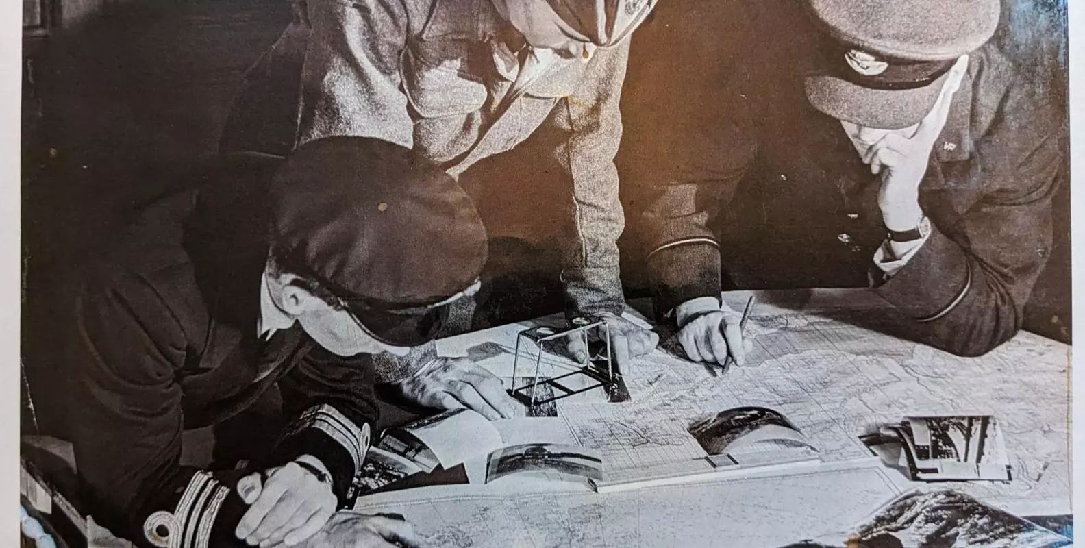 A black and white photograph of three men in military uniforms leaning over photographs and maps 