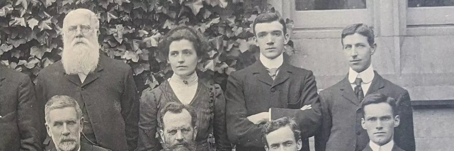 A detail of a black and white photograph showing students posing for a College group picture.  A female figure is standing next to a man with a long white beard.