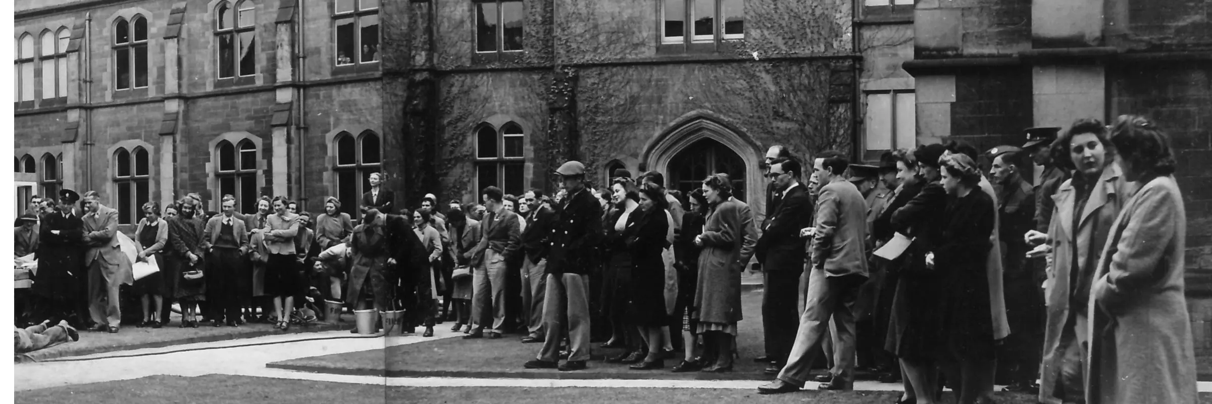 Photograph showing a crowd of people standing in the College quad watching a fire drill beyond the edge of the picture