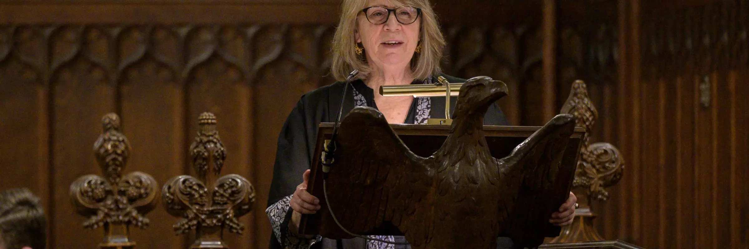 A blonde-hair woman wearing glasses and gown, standing behind the HMC chapel's eagle lectern.